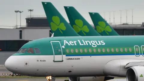 Getty Images Green and white Plane with Aer Lingus written across it with three plane tail wings in the background that each have the Aer Lingus shamrock logo on them