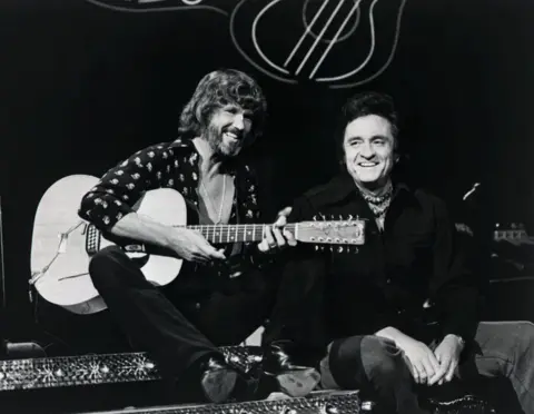 Getty Images Kris Kristofferson, holding a guitar, poses with country music legend Johnny Cash on the set of a TV show in the 1960s.