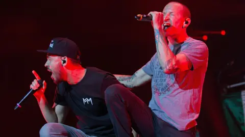 Getty Images Mike Shinoda and Chester Bennington performing in 2015. They both sing, Chester with his hand on Mike's back as they lean forward off the stage. Mile wears a black cap and T-shirt, Chester wears jeans and a grey T-shirt, revealing heavily tattooed arms. 