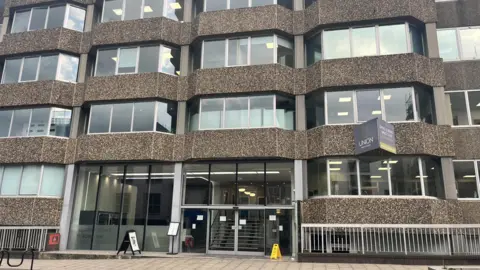 BBC Front aspect of a large office building showing entrance and windows. The pavement can be seen alongside signage. 