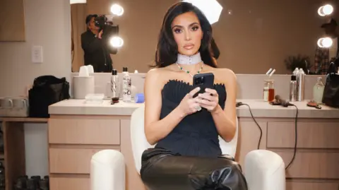 BBC/Passion Pictures/Pierre Auroux/@Pierresnaps Kim Kardashian sitting in a white chair with hair products behind her