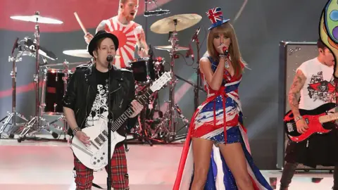 Getty Images Patrick Stump from Fall Out Boy and Taylor Swift stand on a stage at the 2013 Victoria's Secret fashion show. Patrick (left) wears a black fedora, a black ts-shirt, a black leather jacket, red tartan trousers and holds a white guitar. Taylor Swift (right) wears a dress with a union jack on it with a matching cape and top hat. Her microphone is red.