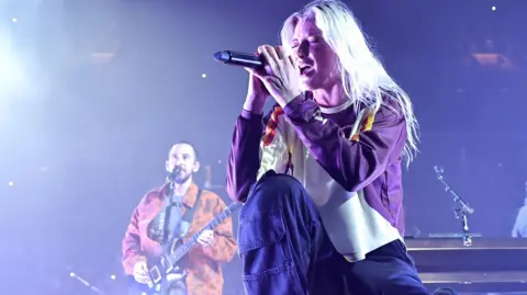 Getty Images Mike Shinoda and Emily Armstrong of Linkin Park perform in New York. Emily, in the foreground, closes her eyes and kneels as she sings into a mic, held in both hands. She has bleached blonde hair worn loose and wears a white and purple sweatshirt. Mike, in the background, plays guitar and sings while wearing an orange jacket. 