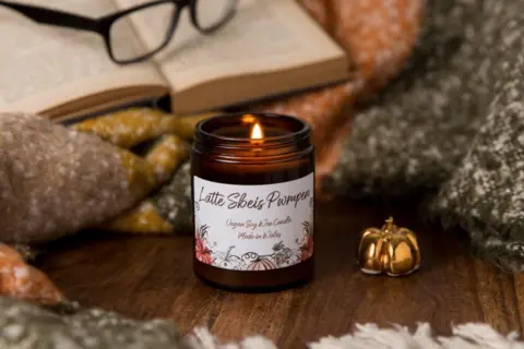 Michelle Huggleston/Little Bit Different A cosy scene of a pumpkin spice candle, on a wooden table in front of a blanket, a book and a pair of glasses. It is next to a gold trinket in the shape of a pumpkin.