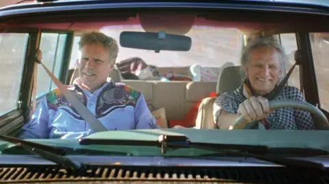 Netflix Will Ferrell and Harper Steele in a car that Harper is driving. He is wearing a blue cowboy shirt and she is wearing a black gingham top