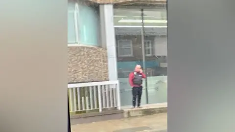 Submitted Blurry picture of a man wearing a tactical vest outside a business premises in Norwich. The man is seen talking on a mobile phone. 