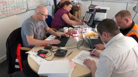 Norsk Hydro Staff at Norsk Hydro crowd around a small desk. Two are working with pens and paper, another is on a laptop.