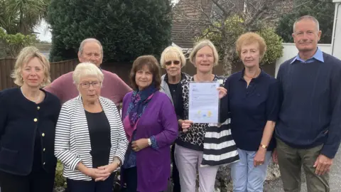 BBC Eight people standing on a surfaced driveway in front of a low brick wall. They are all looking at the camera and one woman is holding a letter on an A4 page up but the contents of the page are too small to read