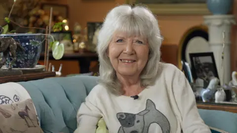 BBC Dame Jilly Cooper sitting on a sofa in the living room of her house in the Cotswolds. She is wearing a dog jumper.