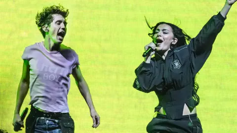 Getty Images Troye Sivan and Charli XCX on stage performing. Troye, a man, is on the left wearing a pink tshirt jumping up, while Charli is in black singing into a microphone with her left arm outstretched. Behind them is a lime green electronic backdrop.