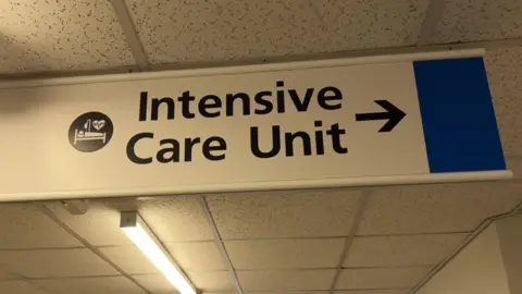 A white sign suspended from a hospital corridor ceiling. 