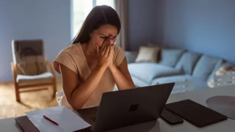 Getty Images Young woman with her face in her hands looking distressed at a laptop screen with her mobile phone, ipad and paper and pen next to her