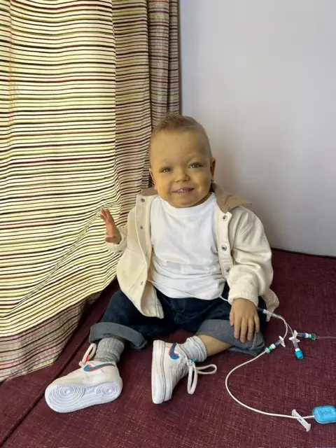 Bradford Family Zachary sits on the carpet beside a stripy curtain, his medical tubes visible beside him