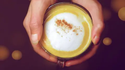 Getty A close up photograph from above of someone's hands wrapped around a glass mug of pumpkin spice latte. It has foamed milk and a sprinkle of cinnamon on top.