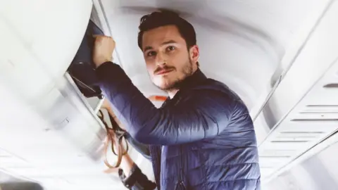 Getty Images Man standing up on a plane with hand on luggage in the overhead compartment