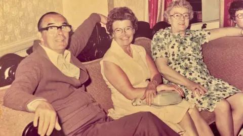 Ian Rankin Jim and Isobel Rankin (middle) smiling at the camera as they sit on a sofa with others in bed and breakfast accommodation