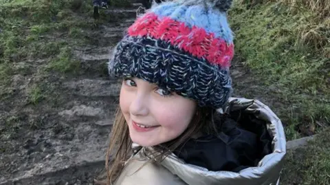 Family handout A little girl smiling wearing a black pink and white beanie hat in a silver puffer jacket