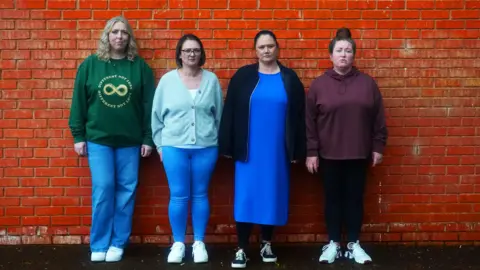 Four women. Far left has blonde hair and is wearing a green sweater and blue jeans, next to her has shot brown hair and is wearing a sky blue cardigan and blue jeans, next to her has brown hair pulled back into a pony tail, a blue dress and darker blue jacket and next to her has brown hair in a top knot, a maroon hoody and dark blue leggings