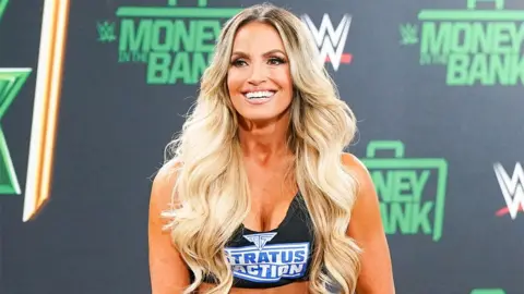 Getty Images Trish Stratus, a woman, on stage at a WWE event for Money In the Bank event, wearing a black top which says 