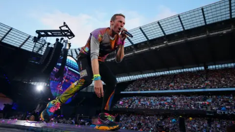 PA Media Coldplay lead singer Chris Martin on stage, wearing fluorescent clothing is bending down and singing. Behind him are thousands of fans in a stadium