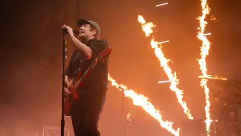 Getty Images Patrick Stump from the band Fall Out Boy is stood on a stage. He stand behind a microphone stand and wears a black shirt, black jeans and a cap. He also has a red guitar. Behind him, fire shoots up from the stage.