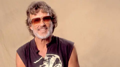 Getty Images Kris Kristofferson poses for a photograph in the 1970s against a beige background, wearing sunglasses and a black sleeveless t-shirt