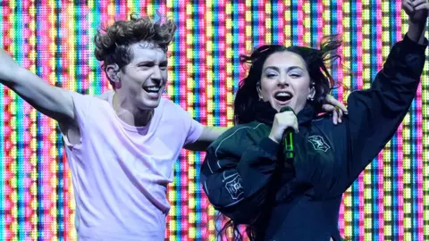 Getty Images Charli XCX and Troye Sivan on stage, with a multi coloured LED lit background. Troye is on the left, wearing a white short sleeved top and has his left arm around Charli with his right arm outstretched. Charli is on the right wearing a black jacket, singing into a black microphone with her left arm outstretched.