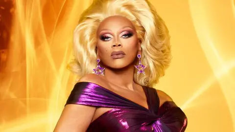 World of Wonder Drag royalty RuPaul is looking directly down the camera lens. She's wearing a dark purple, glittery gown with straps over her shoulders and star-shaped gold and purple earrings. Her eye make-up is dark and dramatic and she has glamorous blonde hair. She's standing in front of a background of billowing orange flames.