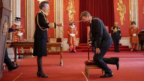 PA Media Ian Rankin kneels on a stool as he is knighted with a sword by Princess Anne  in a palatial room with a red carpet and red and gold walls. There are royal  members of staff in the background.