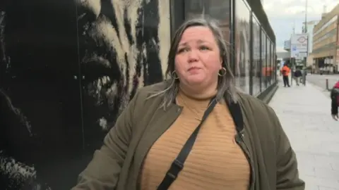 Cassie Smyth, standing on a Birmingham street next to a Peaky Blinders hoarding, is wearing a beige sweat and an olive jacket