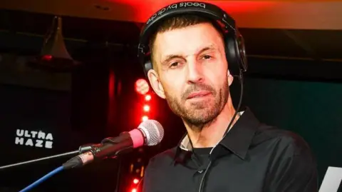 Getty Images Tim Westwood, with a black shirt on, wearing headphones with a microphone in front of him - there are red studio lights behind him and a black backdrop with the words 