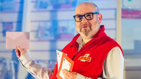 Andrew Billington A cast member in a red Post Office uniform holding up several letters