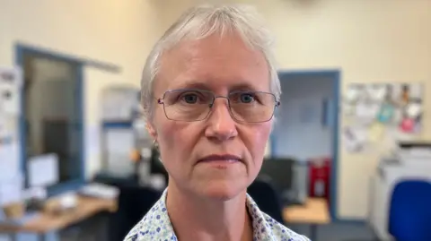 Sam Read/BBC Pat Coomber-Wood is looking at the camera in an office with a straight face while wearing a top with a flower design on it.