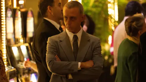 Studio Canal Jeremy Strong as Roy Cohn in The Apprentice - he is surrounded by slot machines and is wearing a grey suit and has his arms crossed