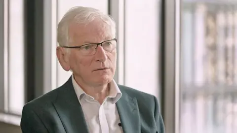 Head and shoulders shot of Dr Dewi Evans, a prosecution medical witness - a man with grey hair, glasses, a grey suit jacket and a white shirt.