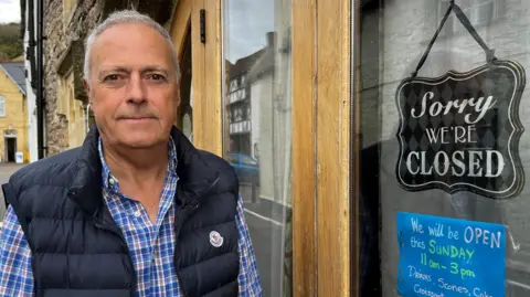 BBC Steve Corrick, a grey haired man in a Moncler-branded functional gilet over a blue checked shirt. He stands outside his teashop in Axbridge town square. A sign on the door says 