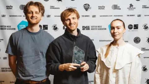 SAY Award Three members of redolent, two male and one female, stand holding their award trophy in front of a wall of sponsor logos