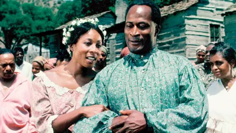 Getty Images Madge Sinclair and John Amos arm in arm in colourful clothes in a scene from Roots in 1977