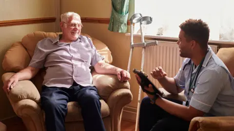 Getty images Older man lounging in armchair with crutches to one side, young male nurse seated in another chair filling in form on a tablet