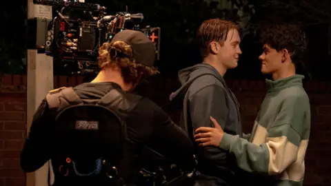 Netflix/Heartstopper Season 3 A camera operator wearing a cap with his back to the photographer films Kit Connor and Joe Locke as they embrace and smile at each other. Kit's ginger hair is wet and he wears a grey hoodie, while Charlie has dark brown curls and wears a baggy green and white jumper.