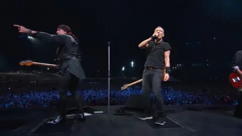 Disney Bruce Springsteen (right) and Steve Van Zandt on stage during his world tour, the pair are both dressed in black and holding guitars. Van Zandt is looking out on the crowd and pointing while Springsteen is looking towards the back of the stage.