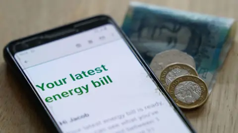 PA Media A phone screen reading 'Your latest energy bill'. A five pound note, two pound coins, and a 50p coin are next to it.