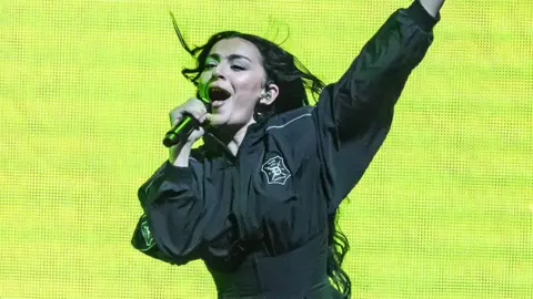 Getty Images Charli XCX, a woman, on stage with a microphone in her right hand, wearing black outfit with the background screen a lime green colour.