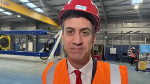 BBC Energy secretary Ed Miliband wearing a red hard hat and orange hi-vis vest, smiling at the camera. 