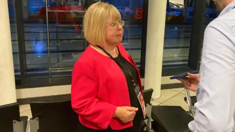 BBC Blaydon and Consett MP Liz Twist. She is being interviewed by a reporter who is holding a phone. Twist is wearing a glasses and a red top. She has blonde hair and is wearing a lanyard.