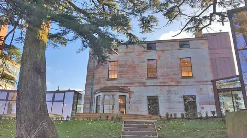 BBC The rear view of a sandstone mansion in Dumfries viewed through a canopy of trees on a clear blue skied day