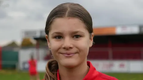 Shaun Whitmore/BBC Isabella faces the camera smiling while standing on a football pitch. She is wearing a red jumper with a collar and her long brown hair has been tied up.