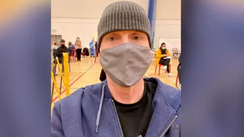 Larry Lowe Larry wearing a grey hat and a grey facemask in a school sports hall which is dotted with people getting their covid jabs