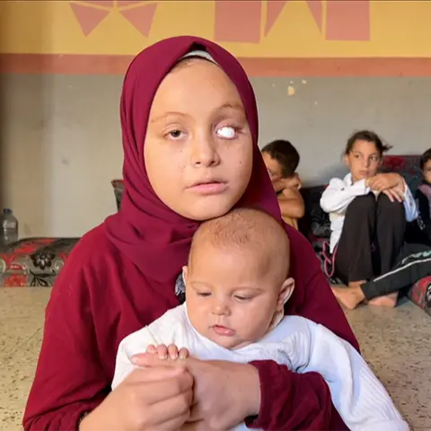 BBC A young teenage girl - Malak - holds her baby sister, Rahma, on her lap. Malak is wearing a burgundy headscarf and long-sleeve tunic. One of her eyes has been replaced with a white sphere. She is looking to the side of the camera. The baby is holding onto her sister's finger and looking at it. She is wearing a long-sleeved white top. In the background, their five siblings sit close together in the corner of a bare room with its walls painted with square patterns,  on large floor cushions. 