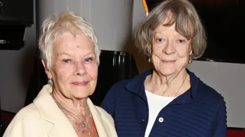 Getty Images Dame Judi Dench, on the left of the image, a woman with cropped white hair wearing an ivory blazer and silver chains, and Dame Maggie Smith, a woman with dark grey hair in a bob wearing a navy jacket over a white top, both smiling